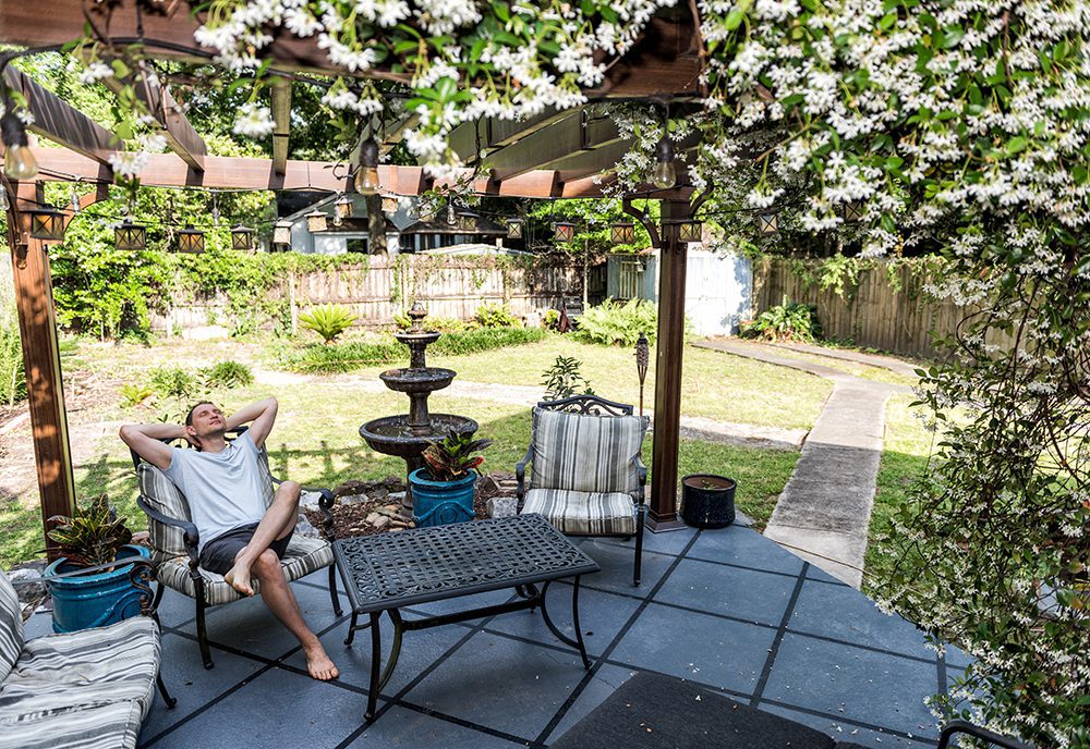 A man relaxing in a chair under a pergola with vining flowers on top of it.