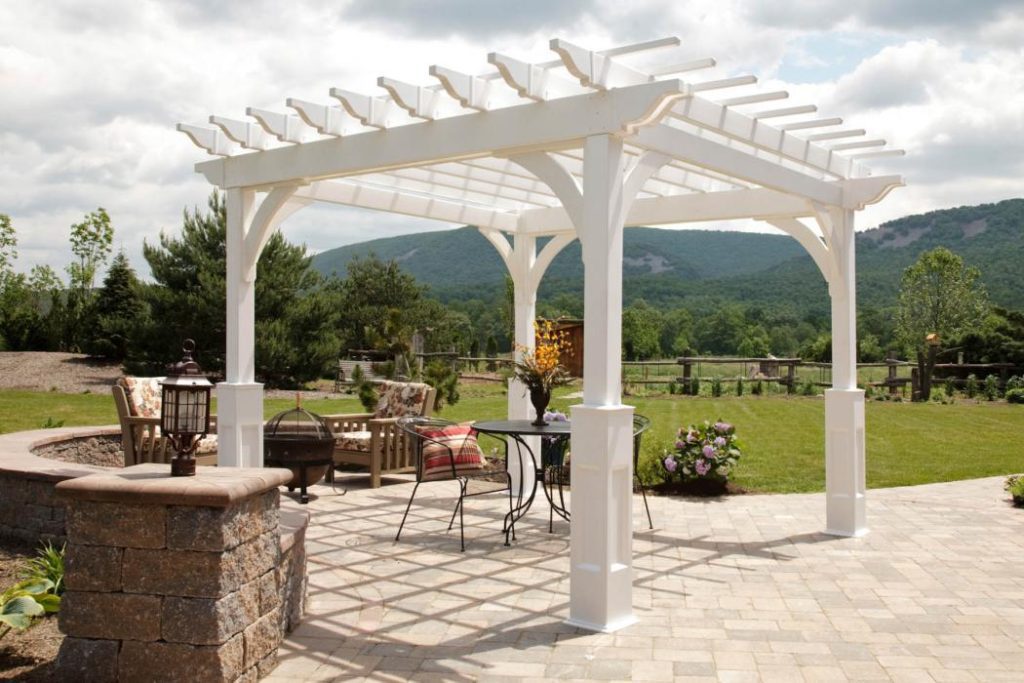 A white pergola on a stone patio with a table and two chairs under it.