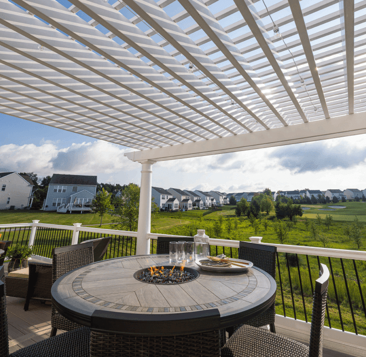 A view from under a pergola with a round fire table on a deck surrounded by chairs.