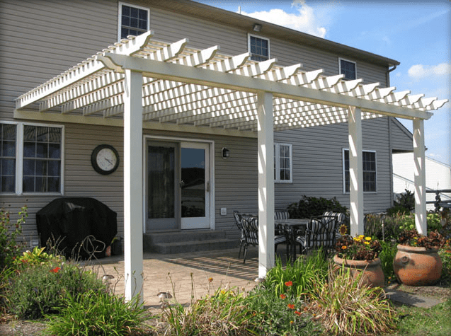 White attached pergola with open slatted roof extending over a backyard patio of a two-story beige house.